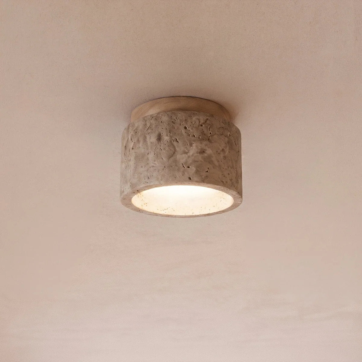 Clérachic compact drum ceiling light in rough travertine with a round wood canopy, its diffuser glowing warm on a ceiling.