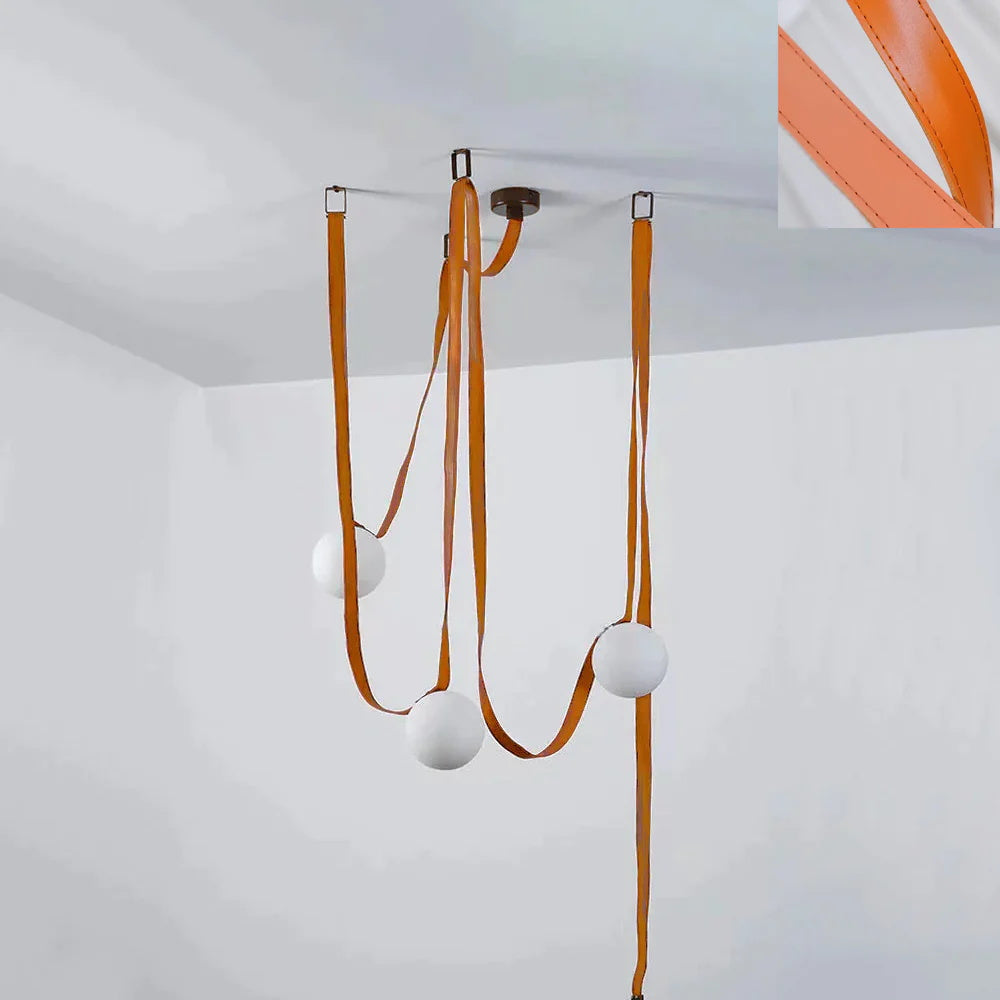Clérachic's Strap-Hung Glass Globe Pendant Light in orange leather, its straps clipped to ceiling hooks with a leather swatch shown alongside.