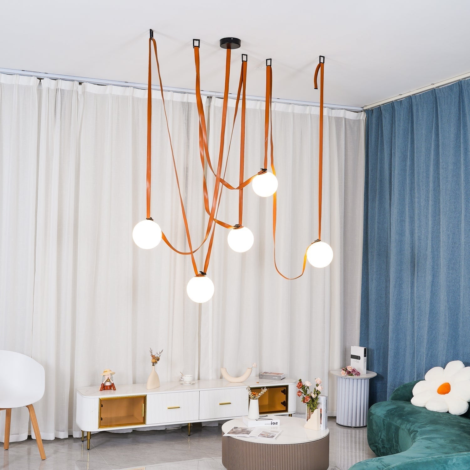 The Clérachic Strap-Hung Glass Globe Pendant Light in orange leather, its five globes staggered above a living room console between sheer and blue curtains.