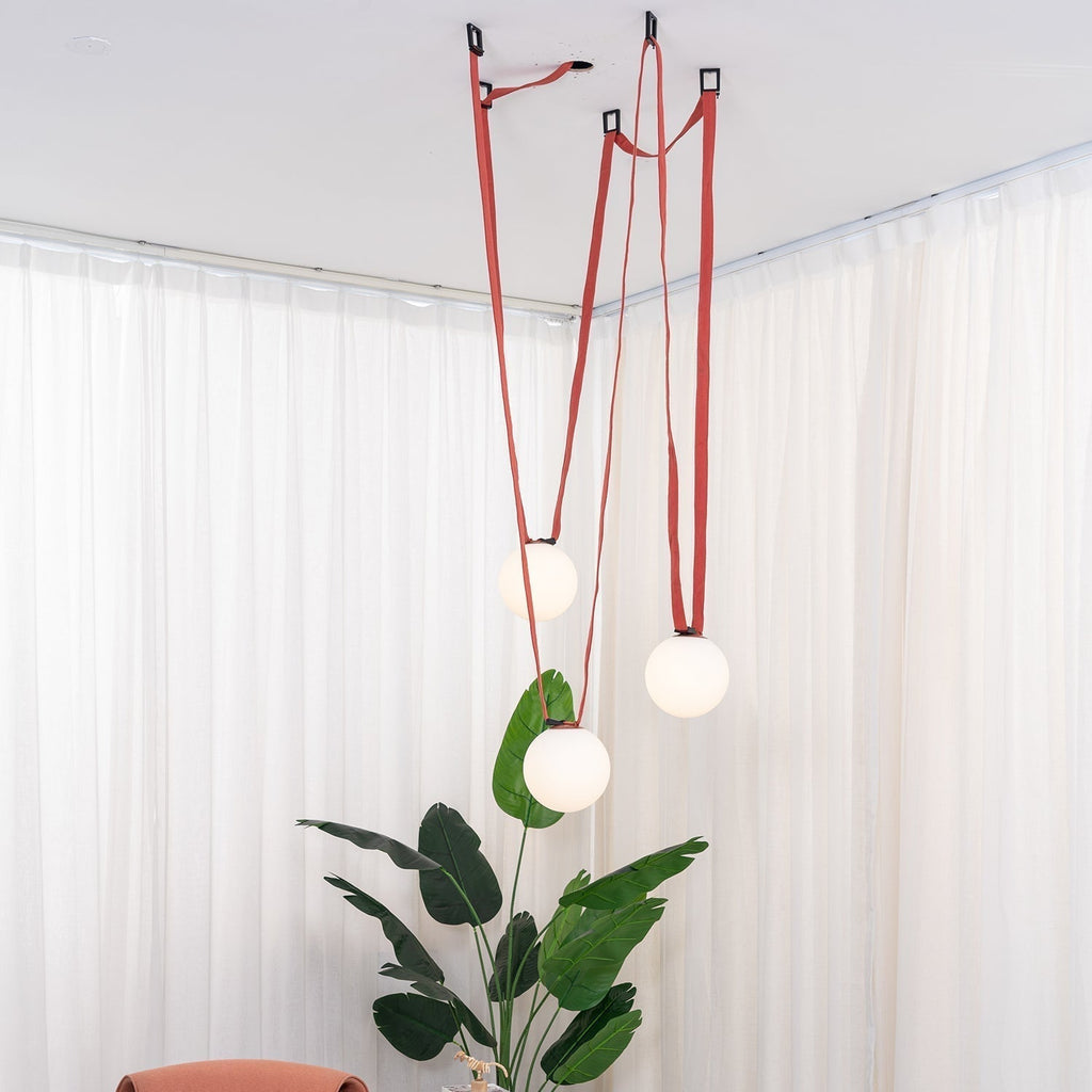 The Clérachic Strap-Hung Glass Globe Pendant Light's red canvas version glowing in a curtained corner, with a large potted plant beneath the lowest globe.