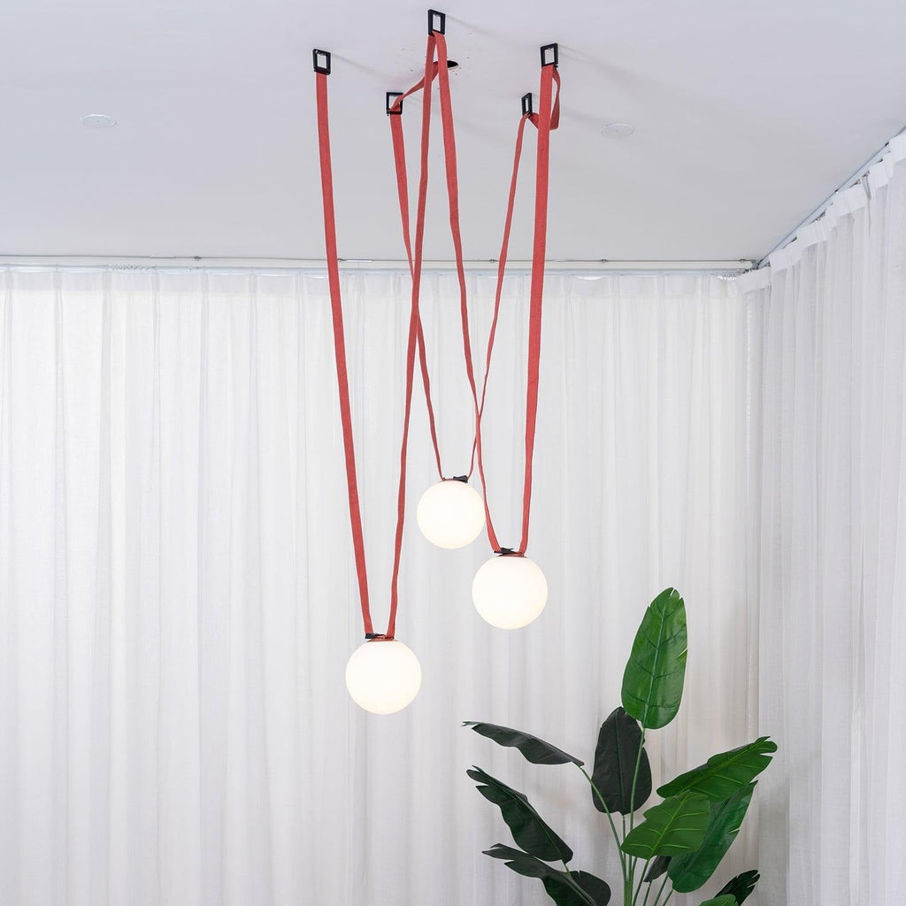 Another angle of the Clérachic Strap-Hung Glass Globe Pendant Light's red canvas globes hanging in front of sheer curtains, with an ottoman edge visible below.