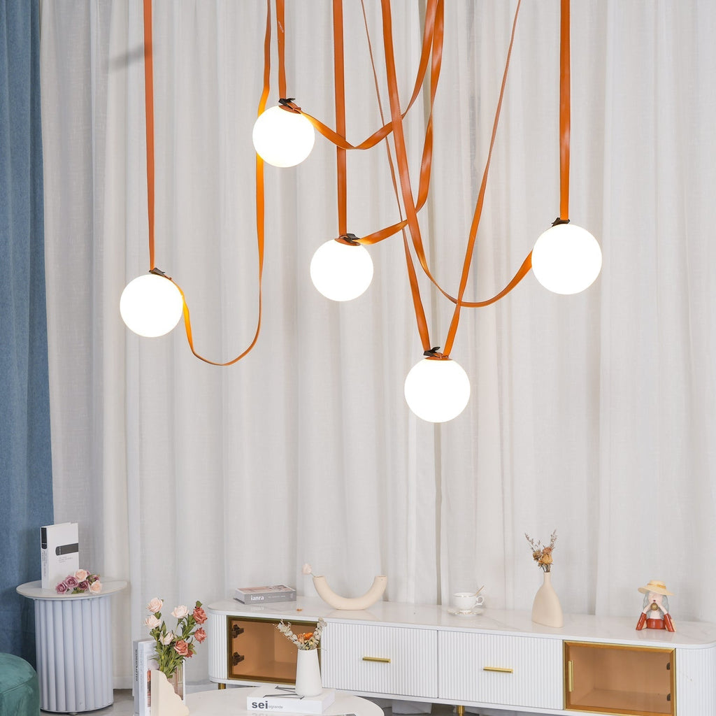 The Clérachic Strap-Hung Glass Globe Pendant Light in orange leather, five globes staggered above a white console table with dried flowers and books.