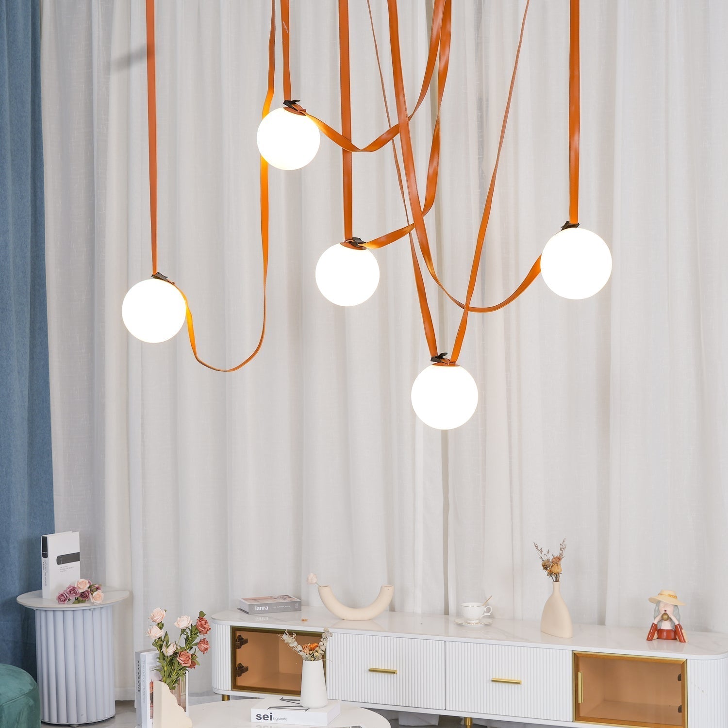 The Clérachic Strap-Hung Glass Globe Pendant Light in orange leather, five globes staggered above a white console table with dried flowers and books.