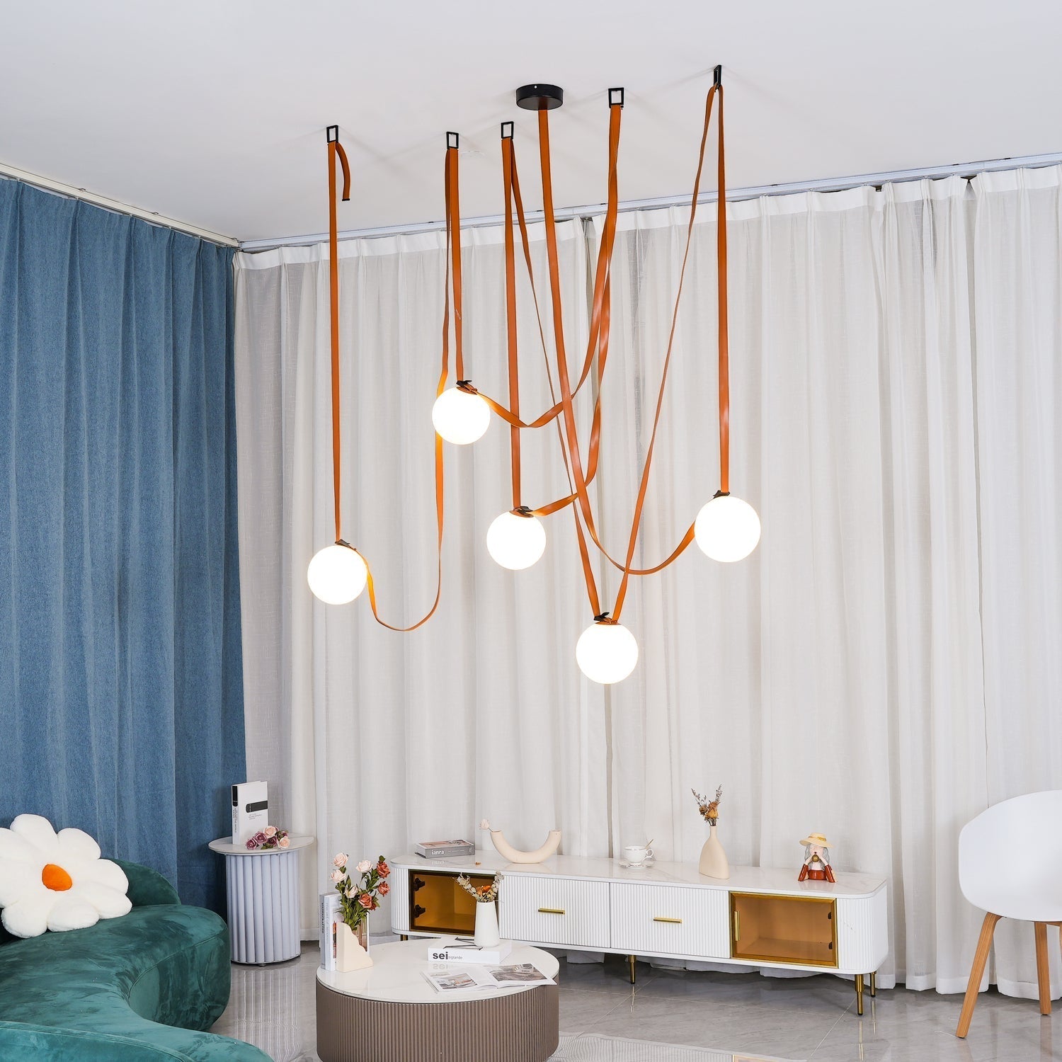Wide living-room view of the Clérachic Strap-Hung Glass Globe Pendant Light in orange leather, its globes glowing between a blue curtain and a green sofa.