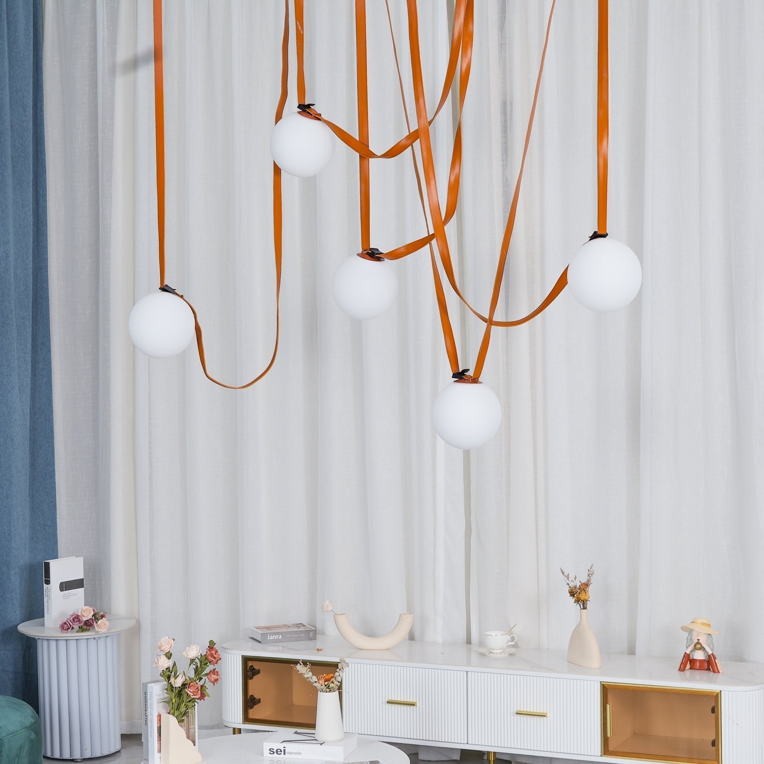 The Clérachic Strap-Hung Glass Globe Pendant Light's orange leather globes staggered over a marble-top console beside a floral arrangement.