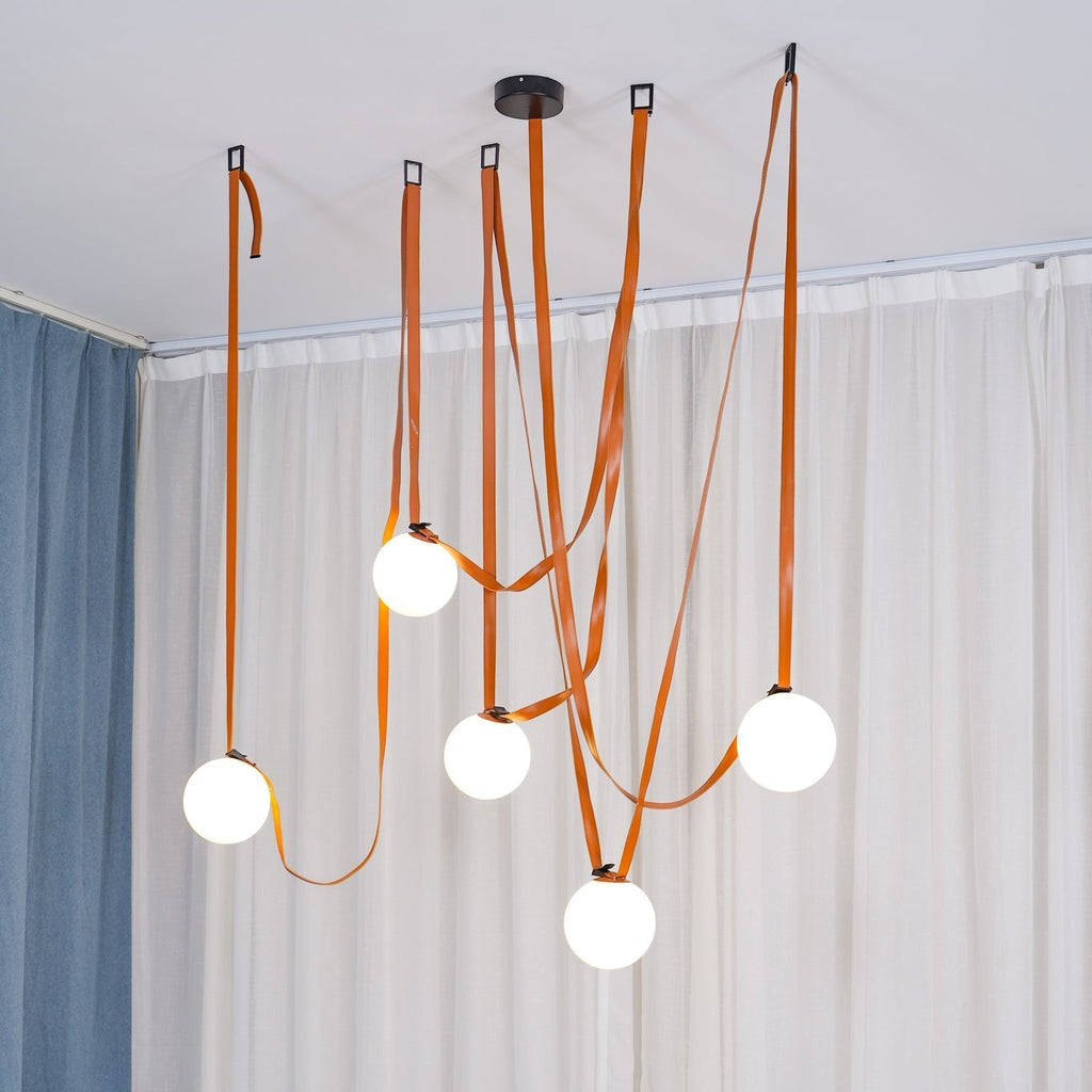 Another full-room angle of the Clérachic Strap-Hung Glass Globe Pendant Light in orange leather, straps crossing above a curtained living area.