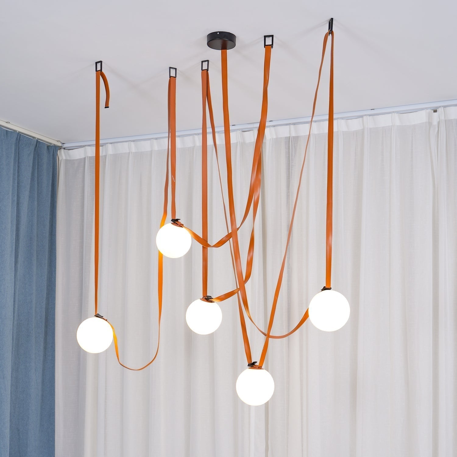 Another full-room angle of the Clérachic Strap-Hung Glass Globe Pendant Light in orange leather, straps crossing above a curtained living area.