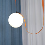 Close-up of a lit globe on the Clérachic Strap-Hung Glass Globe Pendant Light, its leather strap looping between a blue and a sheer white curtain.