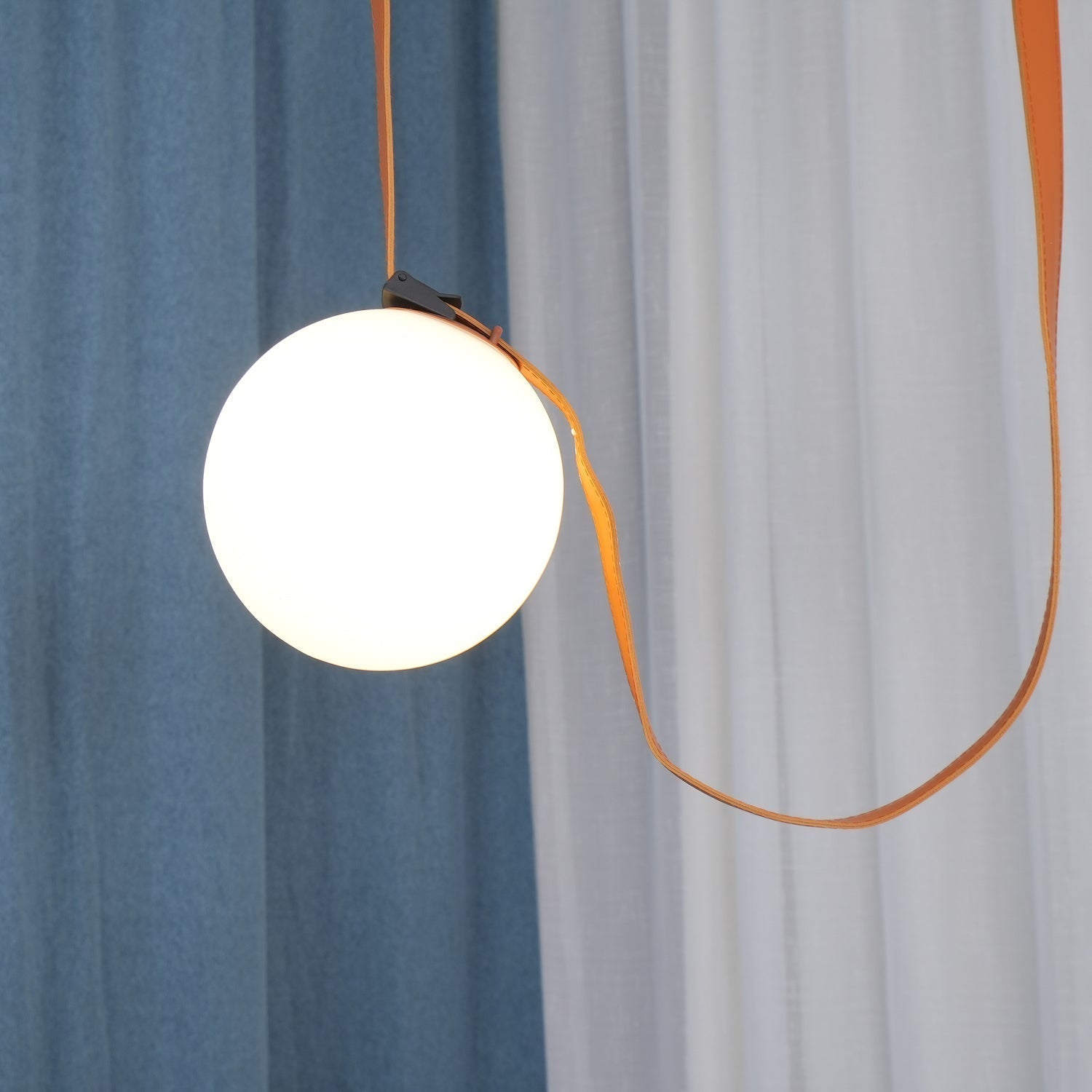 Close-up of a lit globe on the Clérachic Strap-Hung Glass Globe Pendant Light, its leather strap looping between a blue and a sheer white curtain.