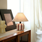 Lit Clérachic table lamp with a red and white striped base on a wooden sideboard, beside a framed print and sheer curtains.