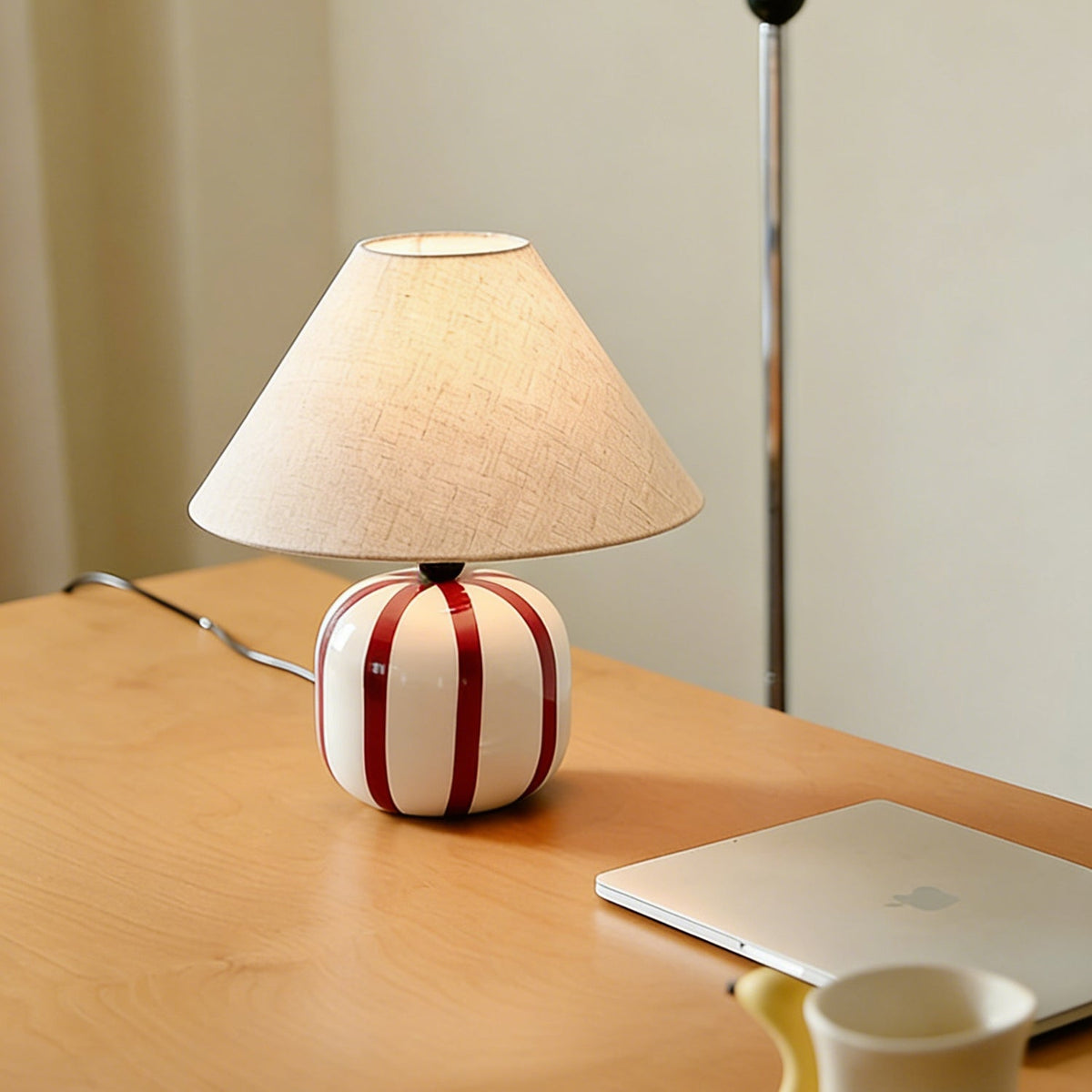 Red and white striped ceramic lamp from Clérachic glowing on a light wood desk, with a laptop and a yellow mug nearby.