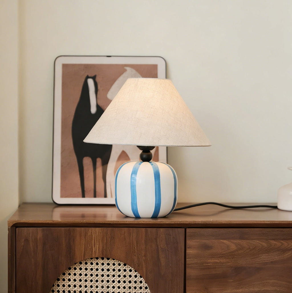 Blue and white striped Clérachic table lamp on a walnut cabinet with a rattan front, in front of a framed horse print.