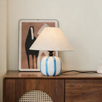 Blue and white striped Clérachic table lamp on a walnut cabinet with a rattan front, in front of a framed horse print.