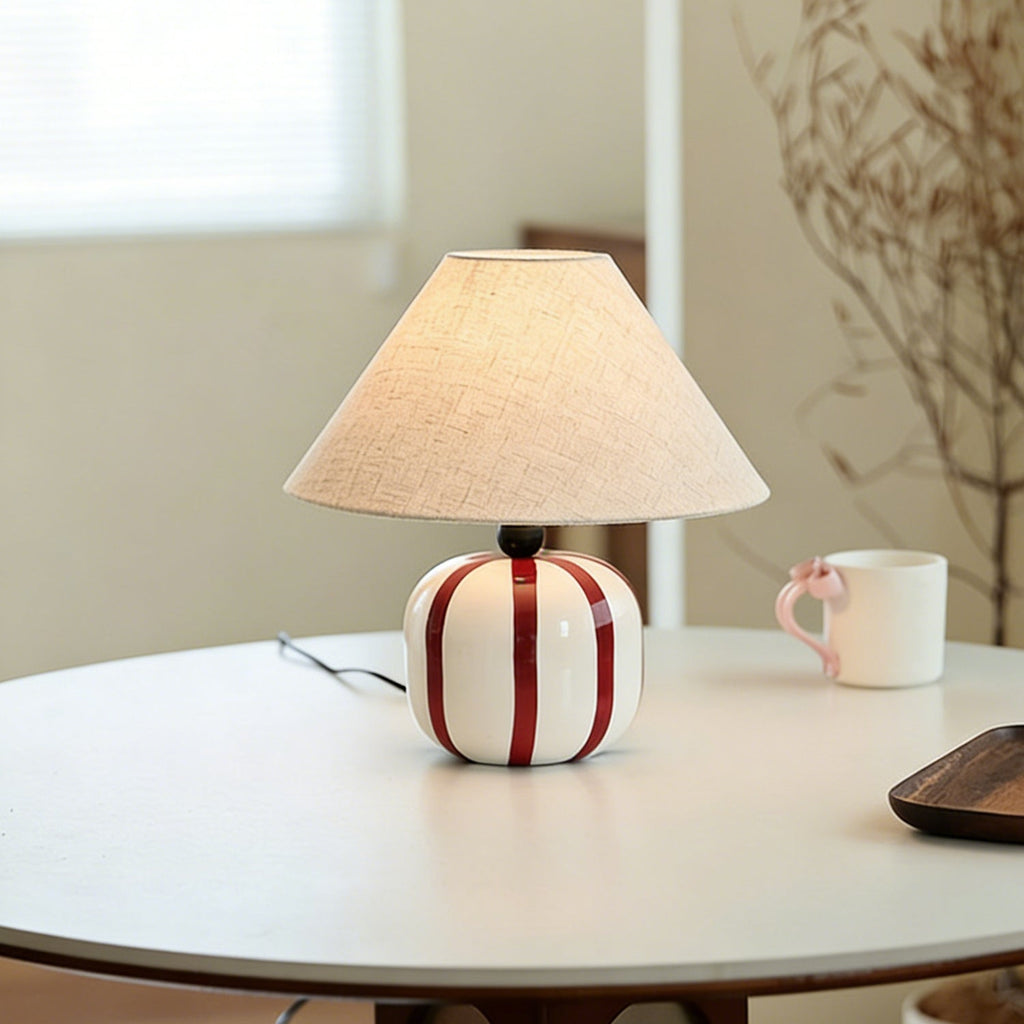 A Clérachic table lamp with red and white stripes on a round white table, a pink-handled mug and dried stems behind it.