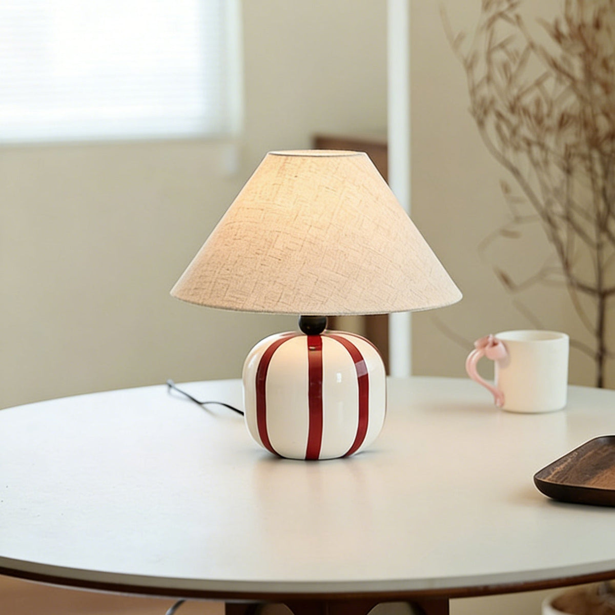A Clérachic table lamp with red and white stripes on a round white table, a pink-handled mug and dried stems behind it.