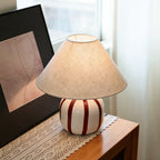 Angled view of a Clérachic lamp with a red and white striped base on a lace runner, next to a dark frame.