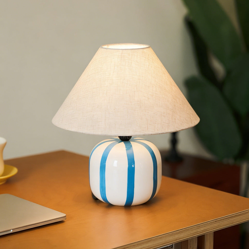 Blue and white striped Clérachic lamp lit on a wooden desk, with a leafy green plant softly out of focus behind it.