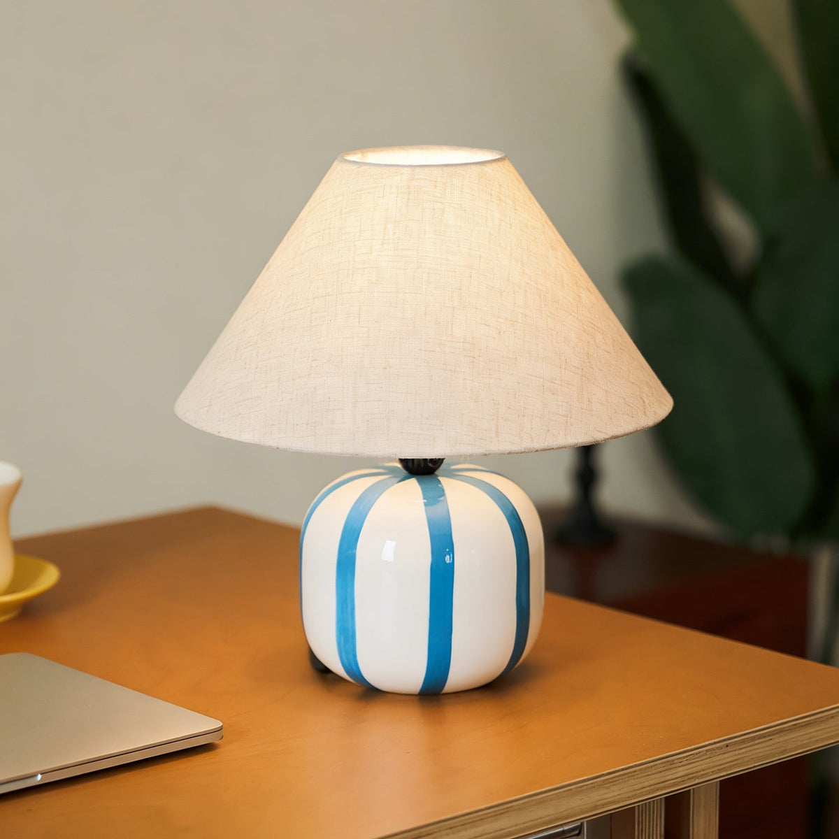 Blue and white striped Clérachic lamp lit on a wooden desk, with a leafy green plant softly out of focus behind it.