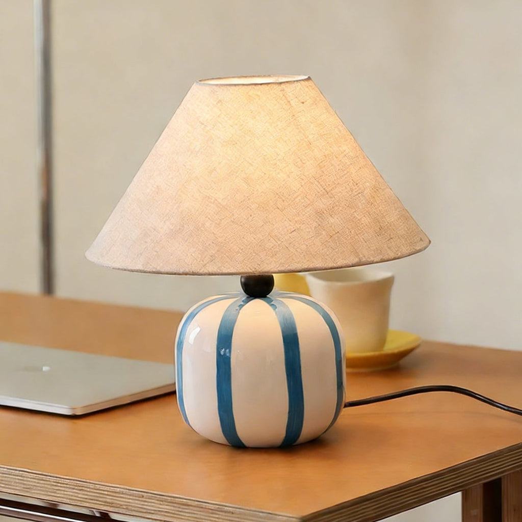 Close front view of a Clérachic table lamp with blue and white stripes, its linen shade glowing above a wooden desk.