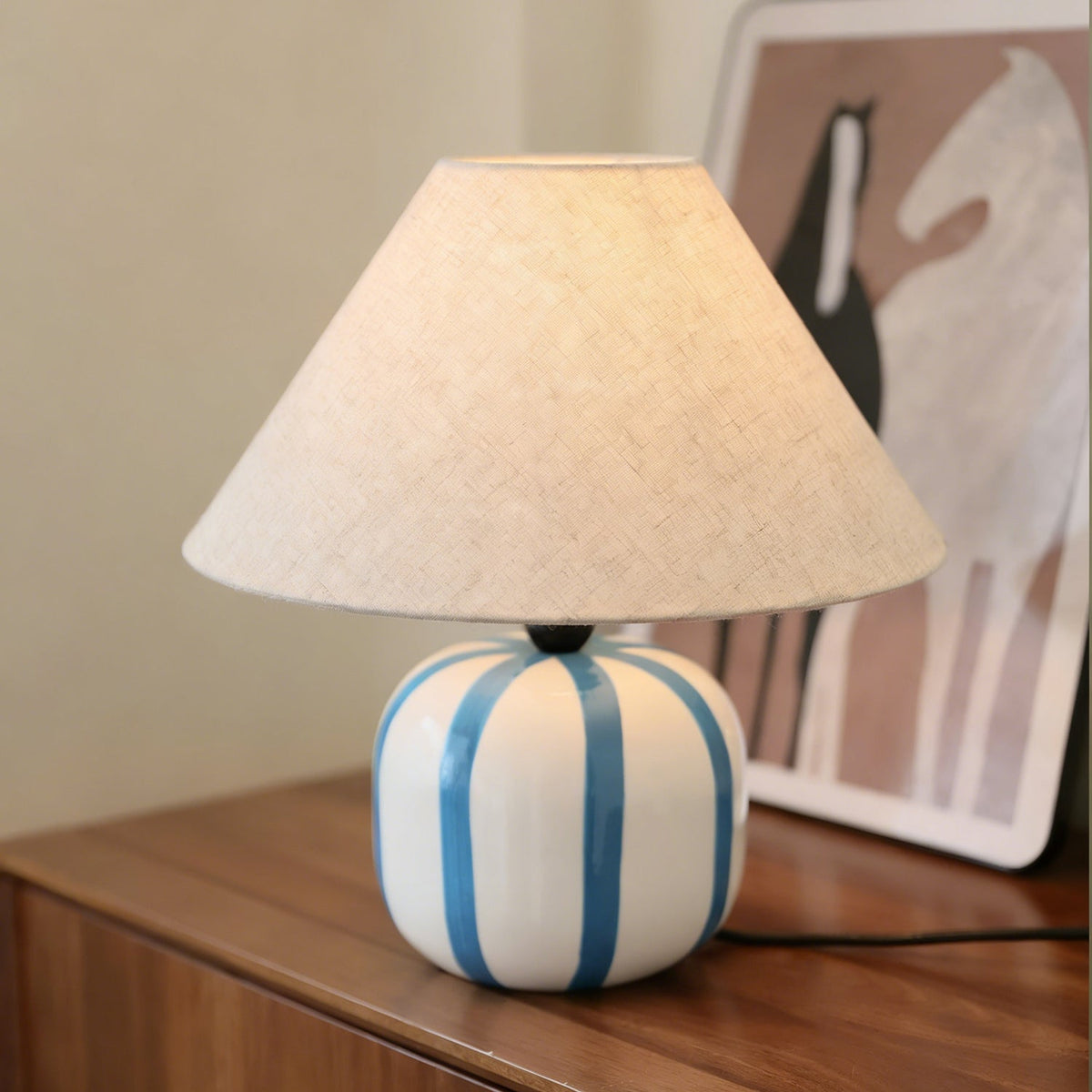 Blue and white striped base of this Clérachic lamp on a dark wood sideboard, with a horse print leaning behind it.
