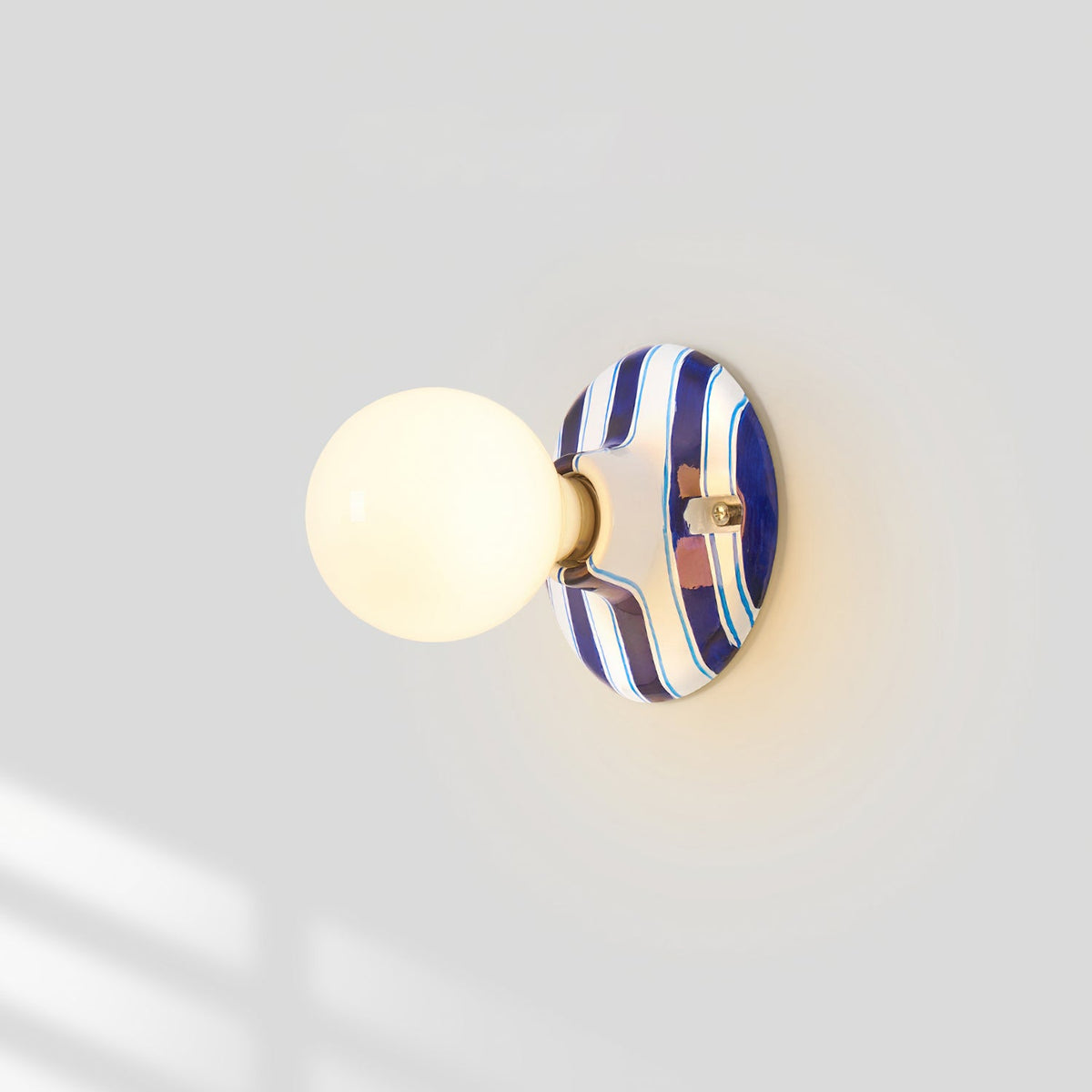 Frosted globe lit on a Clérachic wall light with a blue and white striped ceramic backplate, with soft window light on the wall.