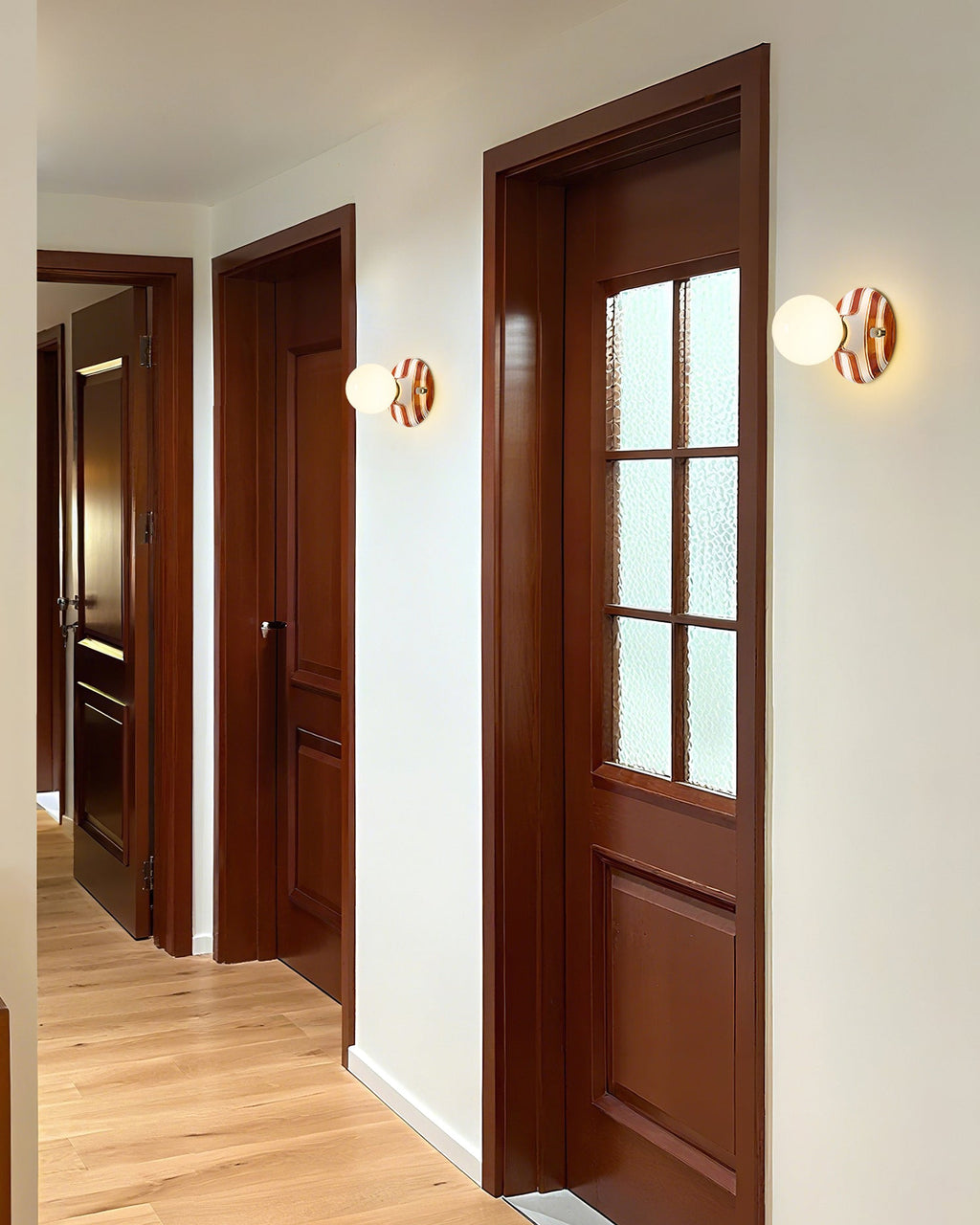 Clérachic wall lights with striped ceramic backplates and frosted globes line a hallway beside dark wood doors and a pale wood floor.