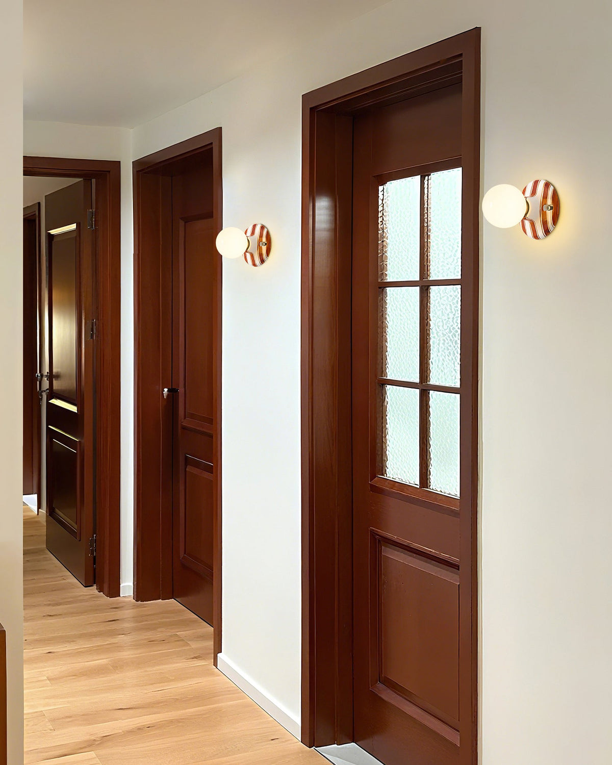 Clérachic wall lights with striped ceramic backplates and frosted globes line a hallway beside dark wood doors and a pale wood floor.