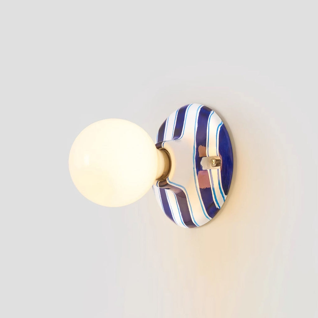 Blue and white striped ceramic backplate and lit frosted globe on a Clérachic wall light, seen from slightly to the side on grey.