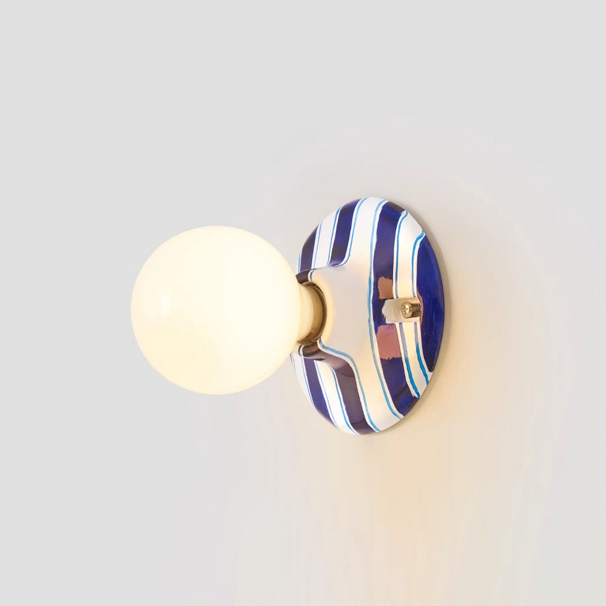 Blue and white striped ceramic backplate and lit frosted globe on a Clérachic wall light, seen from slightly to the side on grey.
