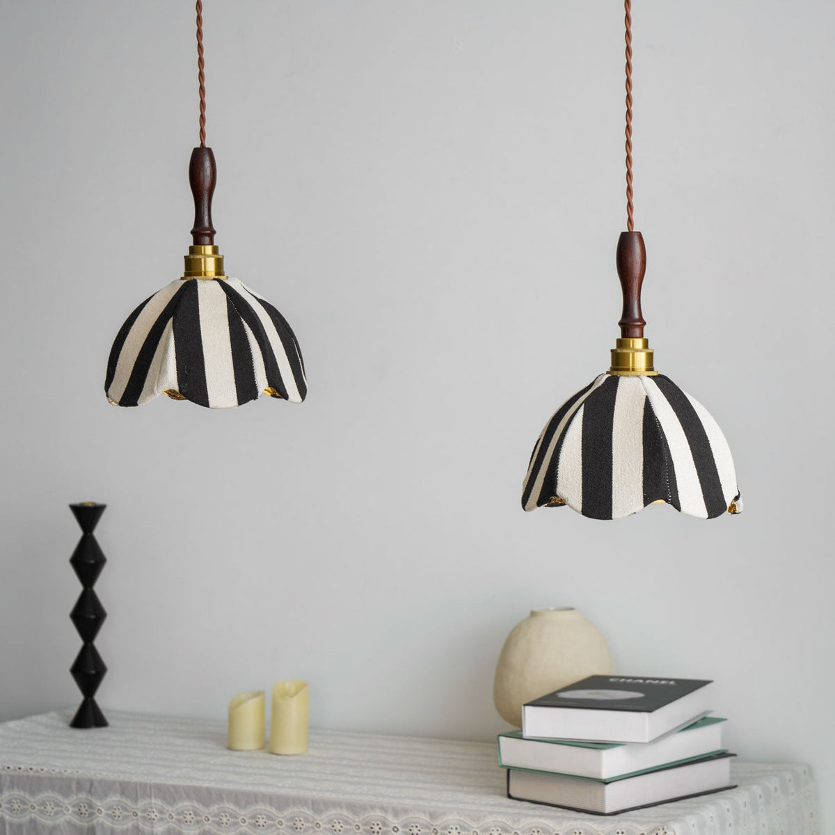 Two Clérachic striped fabric pendants hang above a lace-covered table with books, a pale vase and a black candlestick.