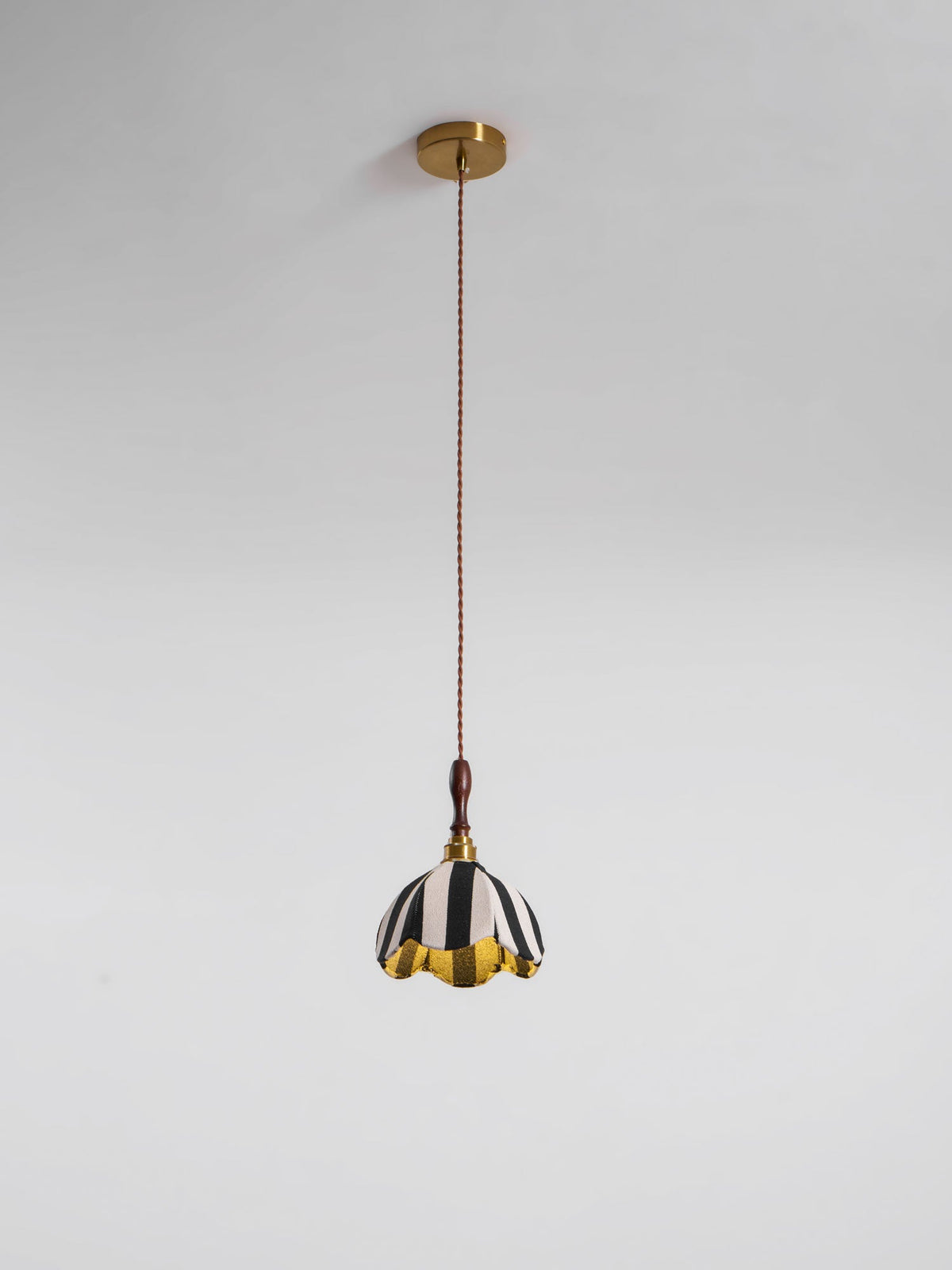 This Clérachic striped pendant hangs from a round brass ceiling canopy on a long brown twisted cord against a grey wall.
