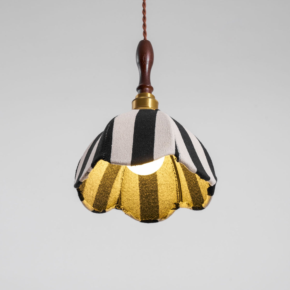 Lit Clérachic striped pendant seen from below, the bulb glowing and the gold lining showing inside the scalloped hem.