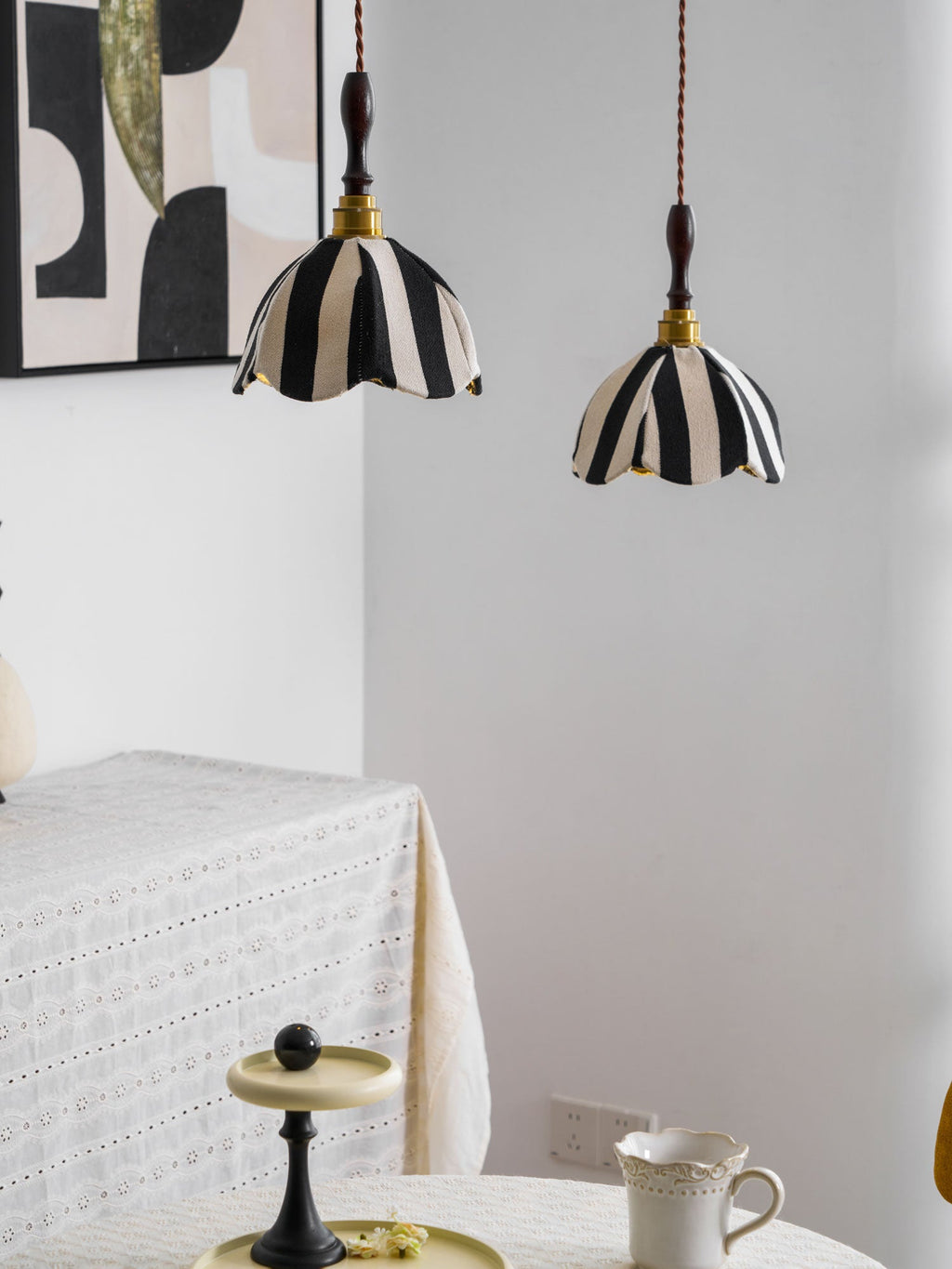 Two striped pendants hang over a corner table with a lace cloth and a teacup, a pair of Clérachic lamps beside a framed print.