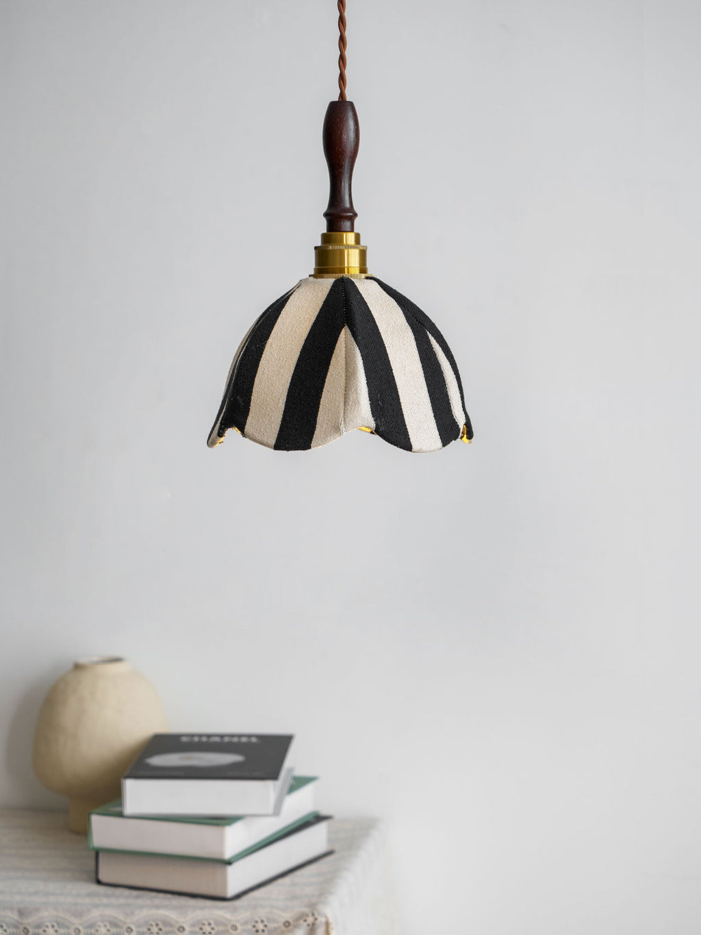 Black-and-cream Clérachic fabric pendant hung above a stack of books and a round stone-coloured vase against a pale wall.