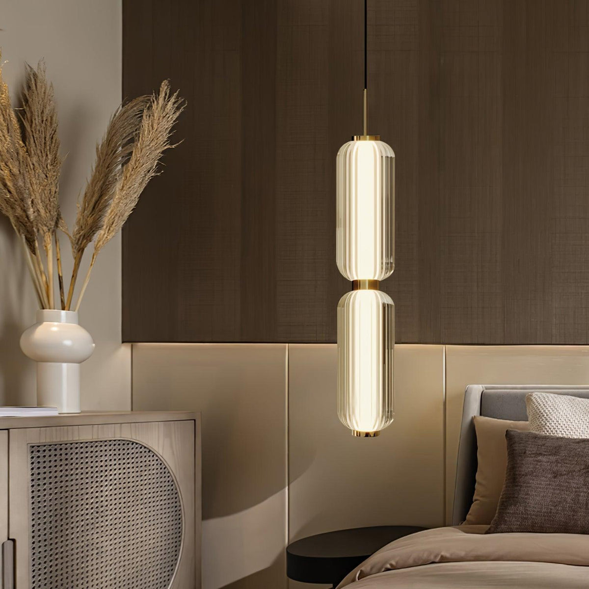 A two-part ribbed glass Clérachic pendant glows beside a bed, with dried pampas grass and a rattan-fronted cabinet behind.