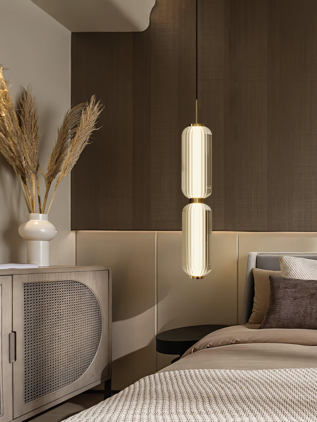 A two-tier ribbed glass Clérachic pendant glowing at a bedside, with dried grasses, a cream vase and a rattan cabinet.