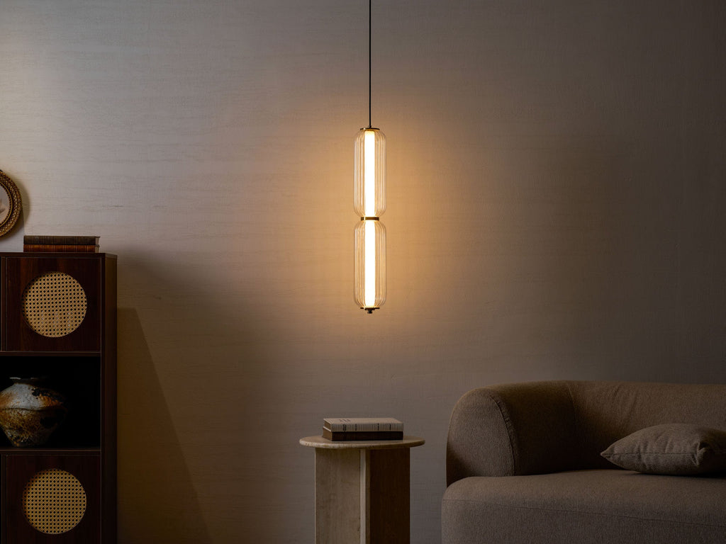 A two-tier Clérachic ribbed glass pendant glows warmly against a dark wall, with a rattan cabinet and a sofa in shadow.