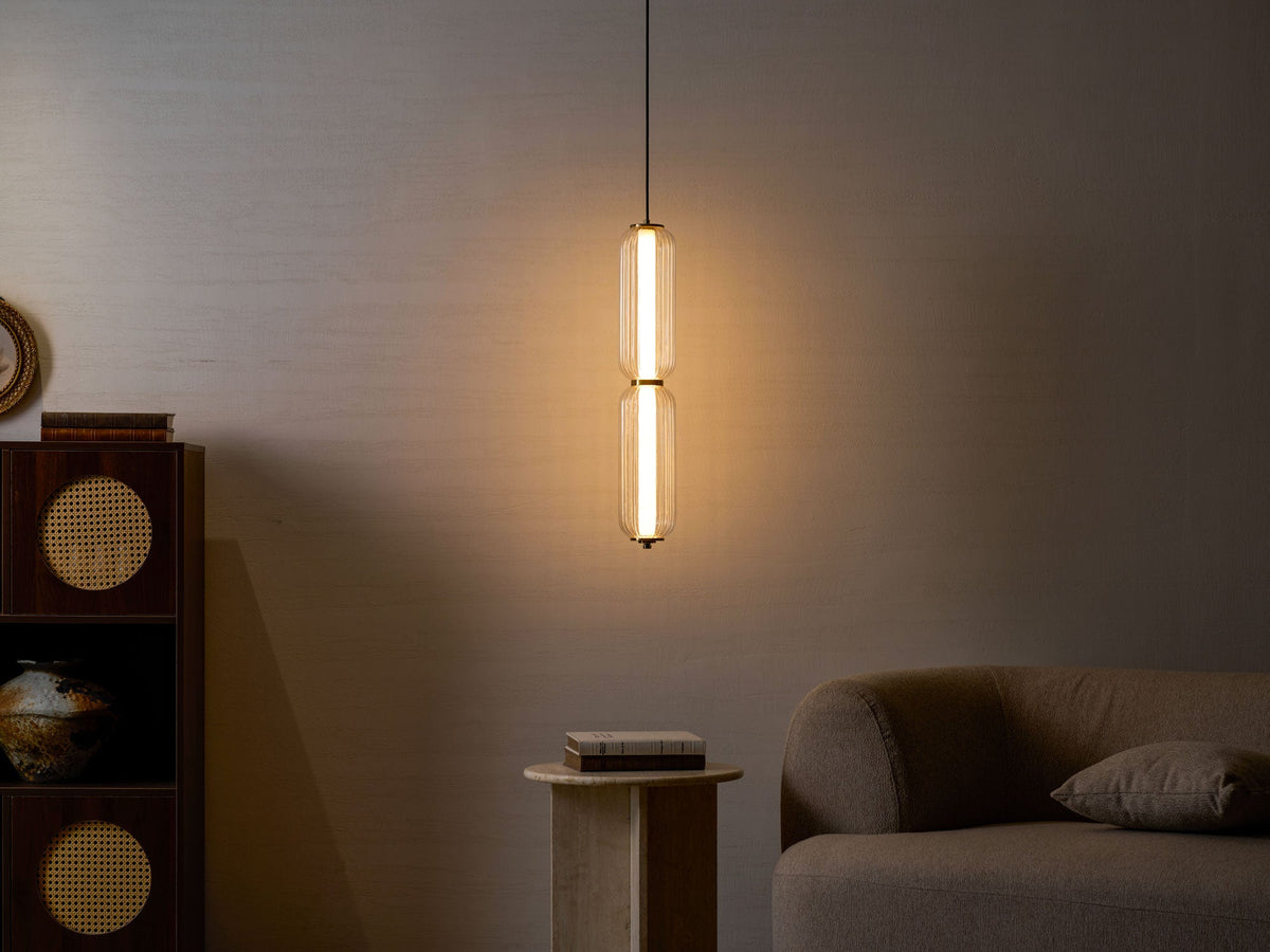 A two-tier Clérachic ribbed glass pendant glows warmly against a dark wall, with a rattan cabinet and a sofa in shadow.