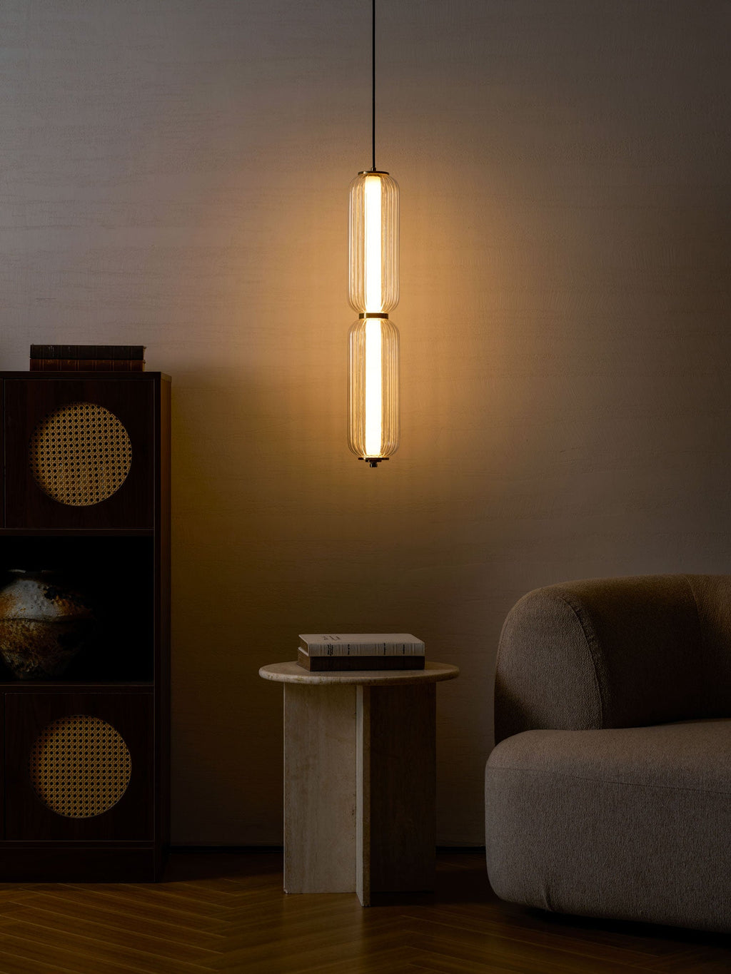 A two-tier Clérachic ribbed glass pendant gives a warm glow in a dim room, above a stone side table, a sofa and a rattan cabinet.