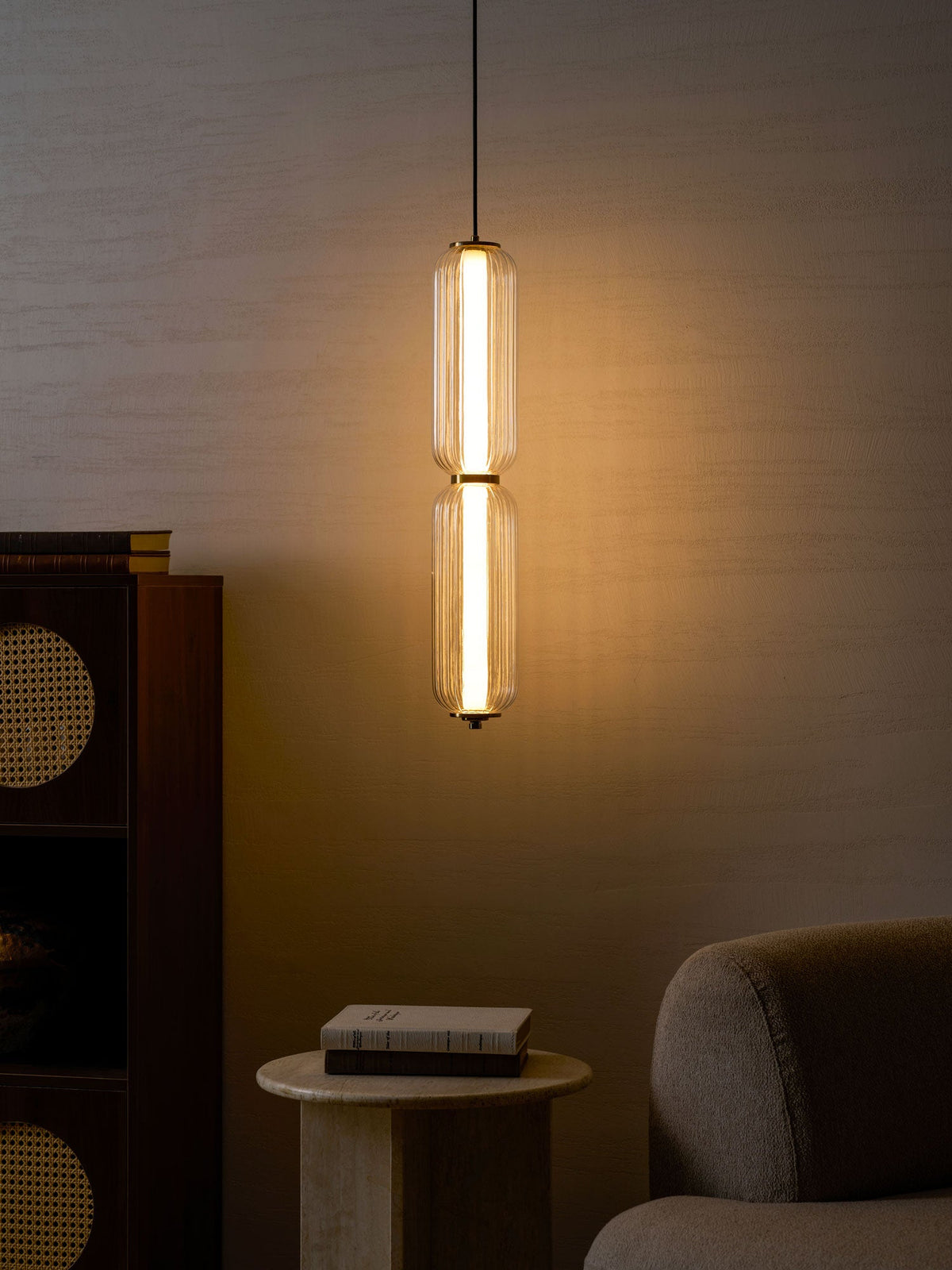 A narrow two-tier Clérachic ribbed glass pendant glows warm gold against a dark textured wall, beside a rattan cabinet.