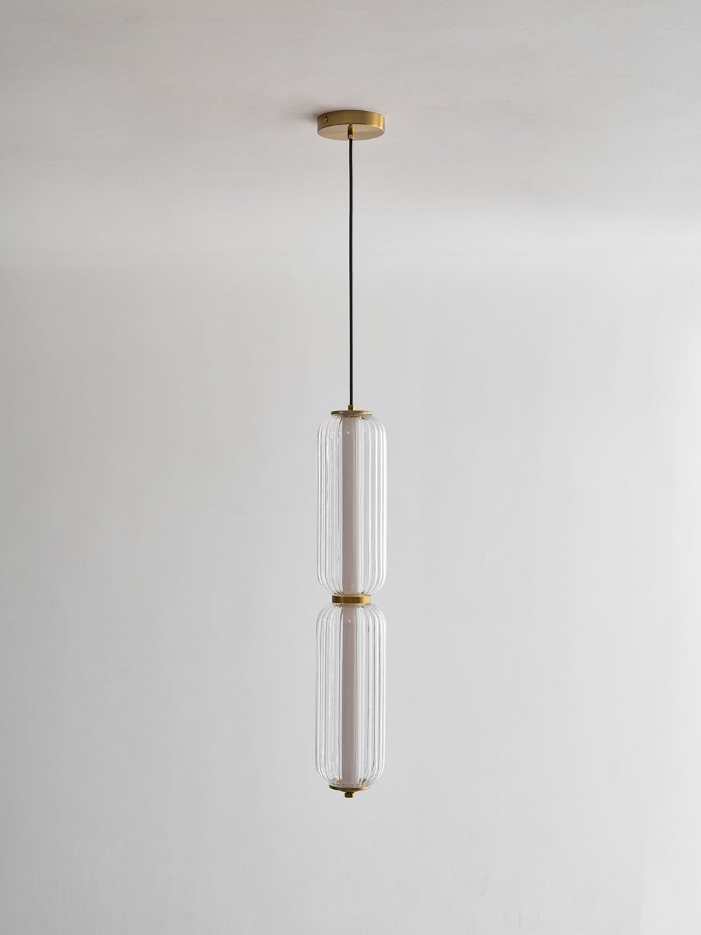 A two-tier Clérachic ribbed glass pendant hangs from a gold canopy on a thin black cord, unlit against a pale wall.