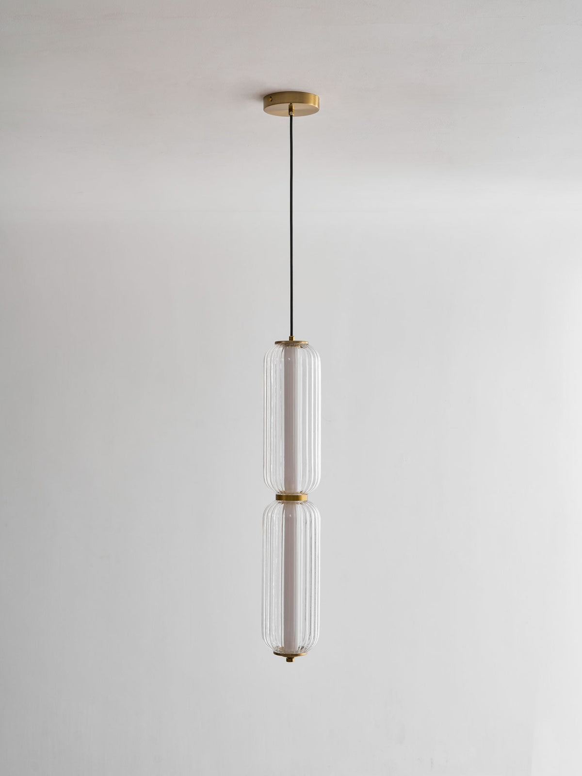 A two-tier Clérachic ribbed glass pendant hangs from a gold canopy on a thin black cord, unlit against a pale wall.