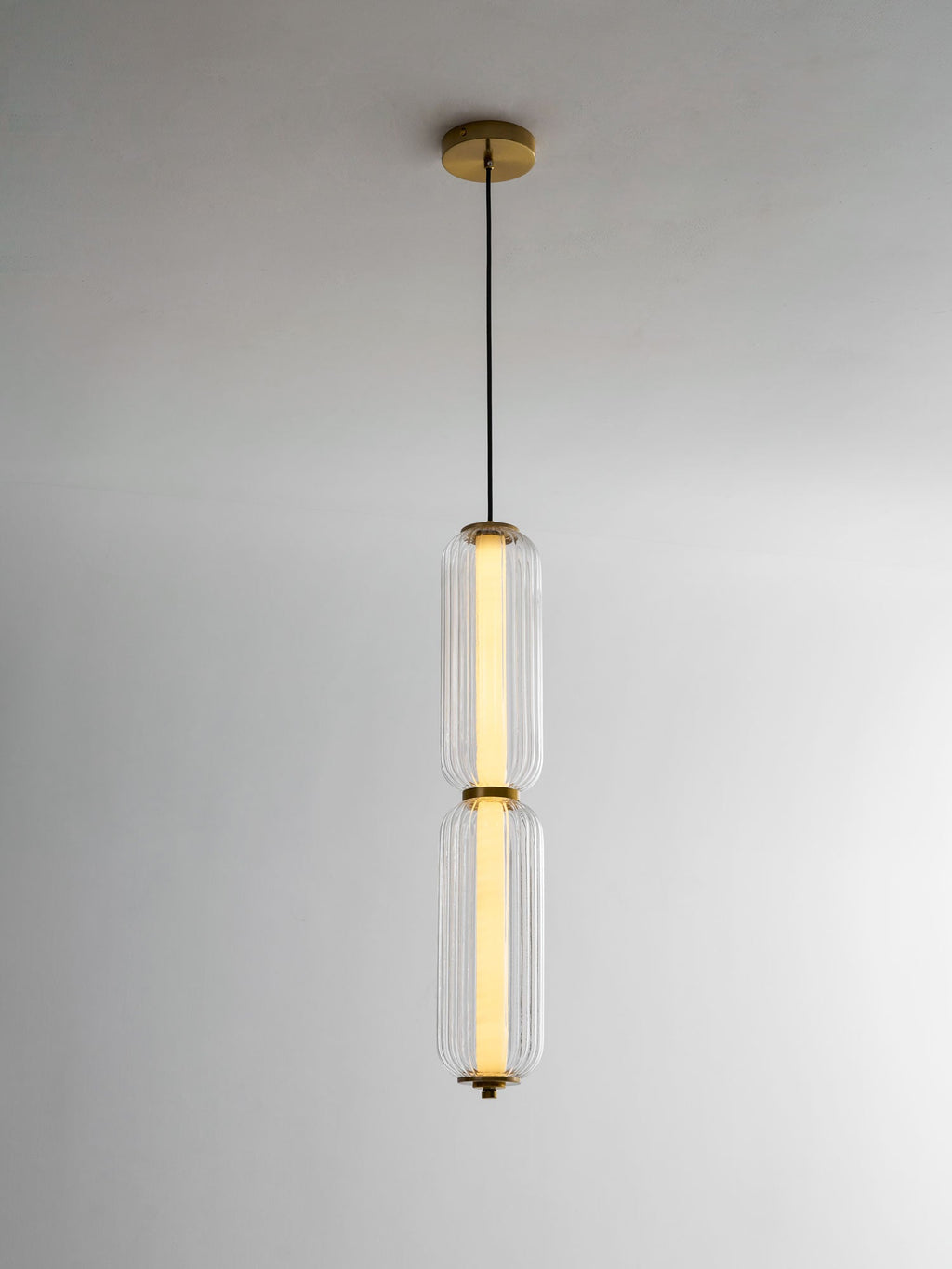 A two-tier ribbed glass Clérachic pendant on a gold canopy, lit warm along its inner tube against a pale wall.