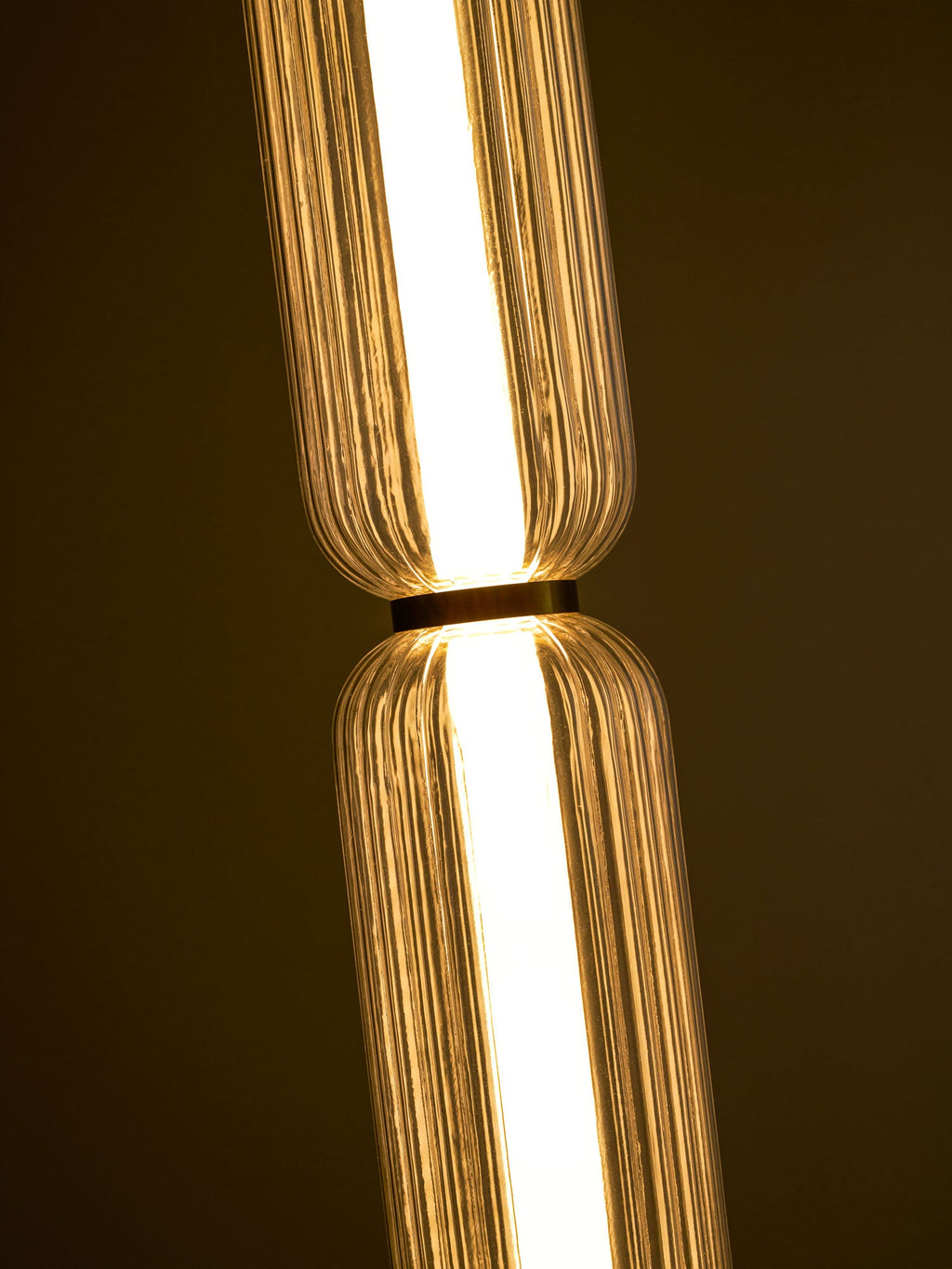 Close view of the gold collar between two lit, ribbed glass tiers of a Clérachic pendant, against a dark background.