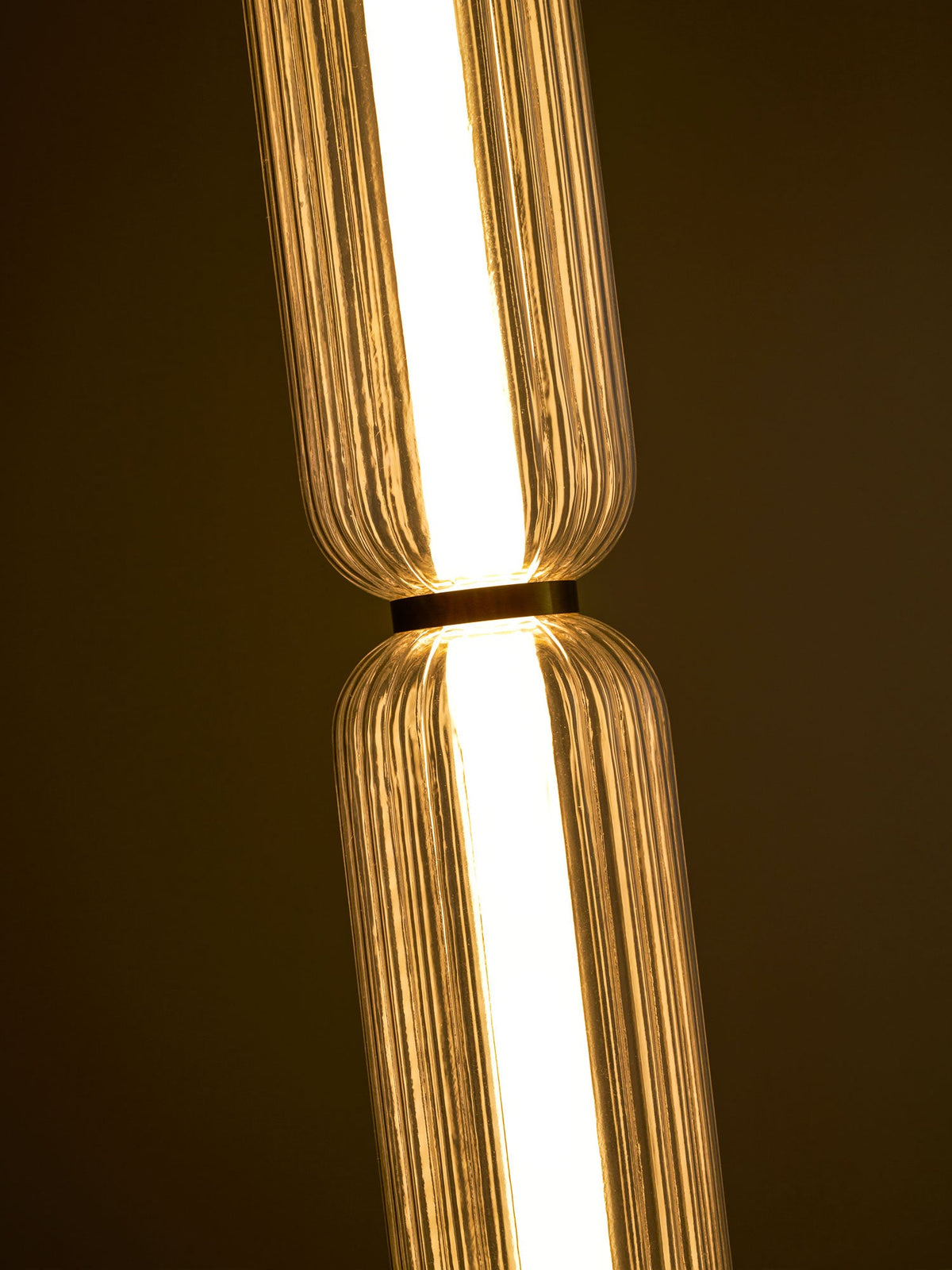 Close view of the gold collar between two lit, ribbed glass tiers of a Clérachic pendant, against a dark background.