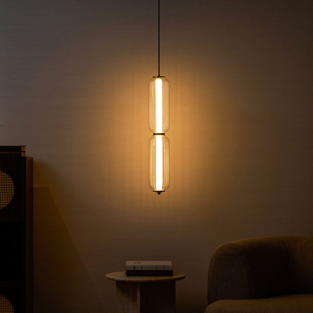 A two-tier Clérachic ribbed glass pendant glows amber in a dim room, with a stone side table, books and a sofa.