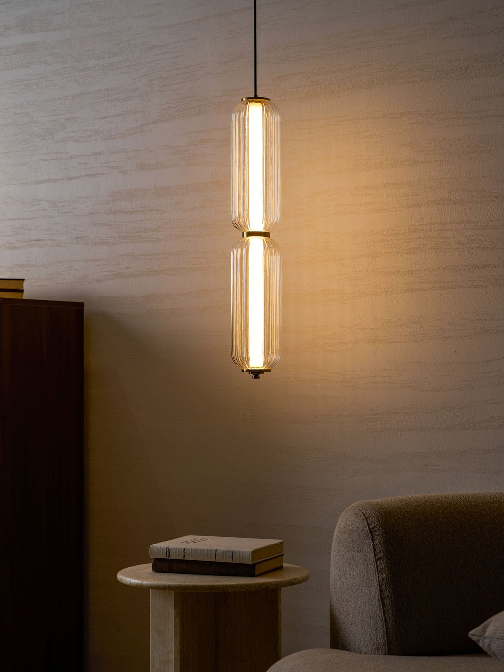 A two-tier ribbed glass Clérachic pendant glows warm in the evening, beside a dark cabinet, a stone table and books.