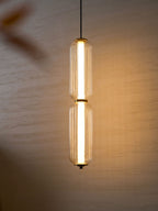 A two-tier ribbed glass Clérachic pendant glows against a textured wall, with a soft blur of an object in the foreground.
