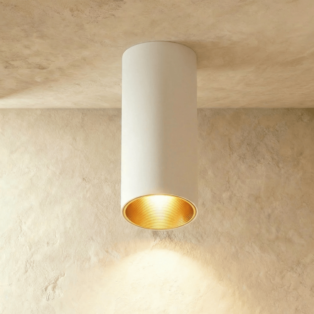 Clérachic surface downlight in white aluminium with a gold-tone baffle, lit warm against a textured plaster ceiling.
