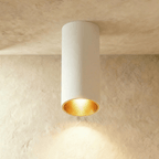 Clérachic surface downlight in white aluminium with a gold-tone baffle, lit warm against a textured plaster ceiling.